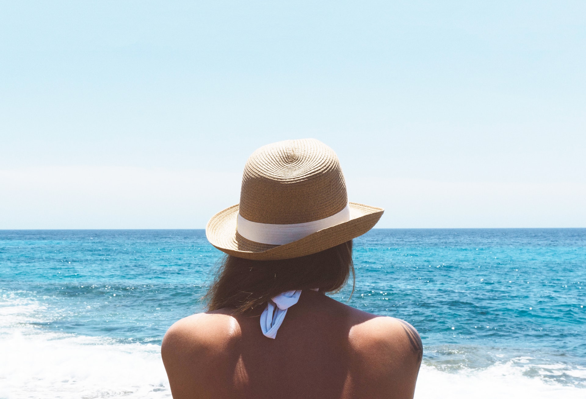 person sits on the beach facing the sea away from the camera, shoulders & back skin exposed, wearing a hat, summer skincare tips