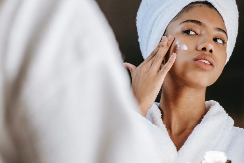 woman with towel on head applying face cream in mirror