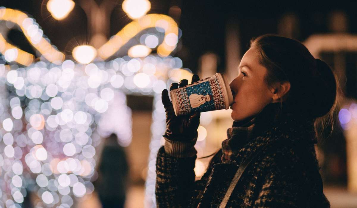 person outside looking happy, drinking from coffee cup, Christmas lights in background