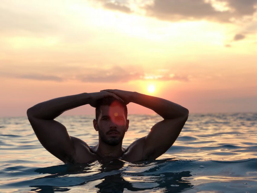 muscled man in water at sunset, hands on head