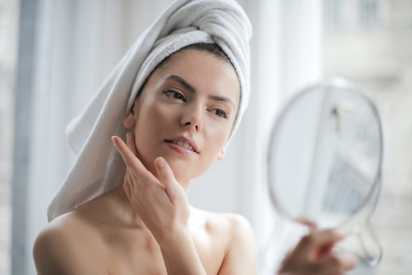 woman with towel on head checks herself in mirror, hand on cheek