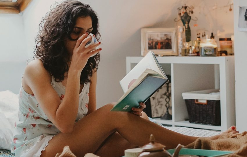 person with healthy looking skin  reading a book while sitting on bed and drinking from cup