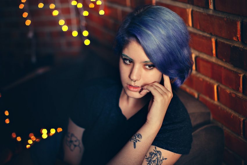 woman with blue hair and tattoos sits pensive on couch, wall and fairy lights in background