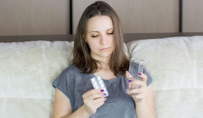 Person sitting on bed holding a glass of water in one hand and painkillers in the other